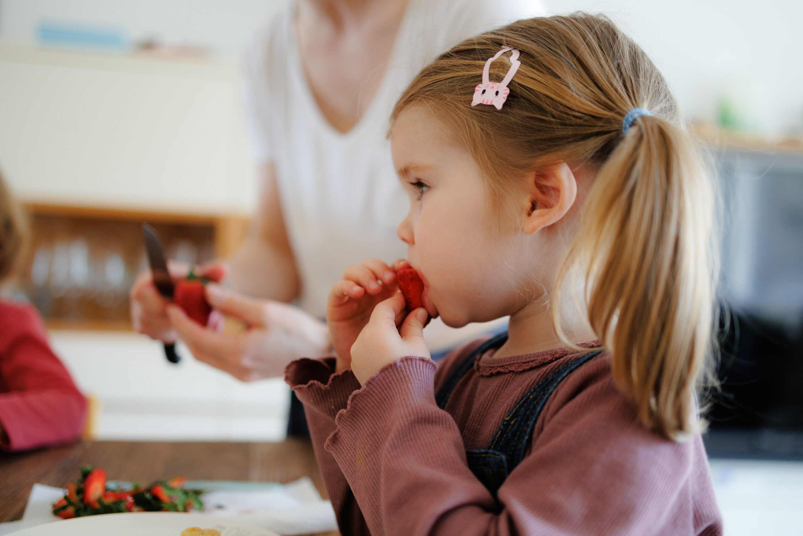 Kleines Mädchen mit Zopf und Haarspange isst während einer Familien-Reportage eine Erdbeere, die Mama im Hintergrund vom Grün befreit.