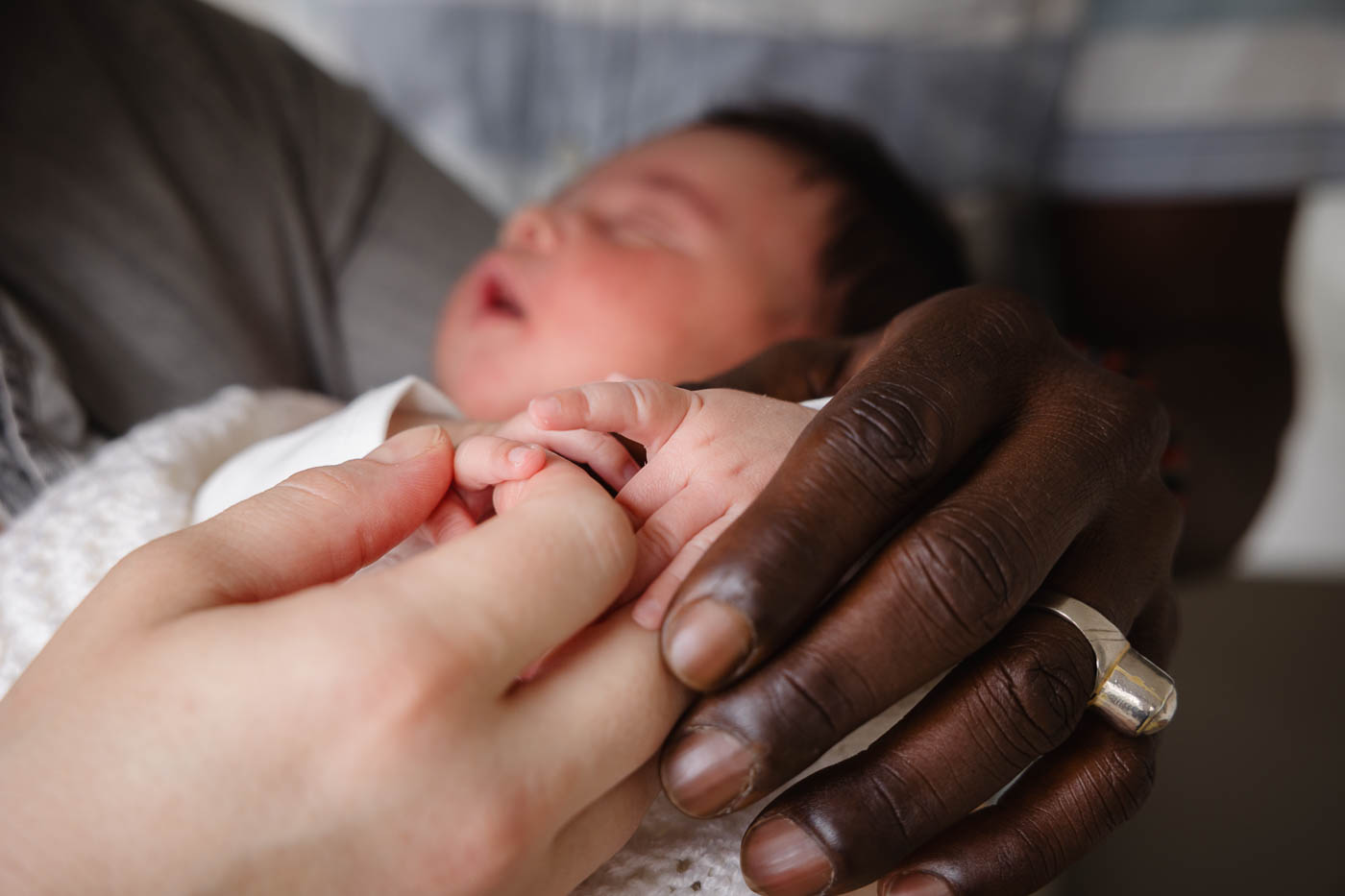 Nahaufnahme der Hände einer Familie bei einem Neugeborenen-Fotoshooting, die Hände der Mama und des Papas halten zärtlich die kleinen Finger des Babys, im Hintergrund sieht man verschwommen das Gesicht des schlafenden Neugeborenen.