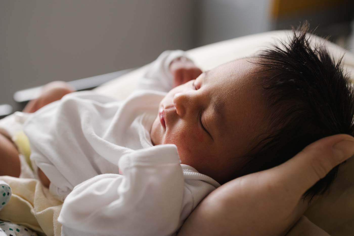Die zarten Gesichtszüge eines schlafenden Neugeborenen im weißen Body, im Krankenhausbett liegend, den Kopf geborgen auf Mamas Hand..
