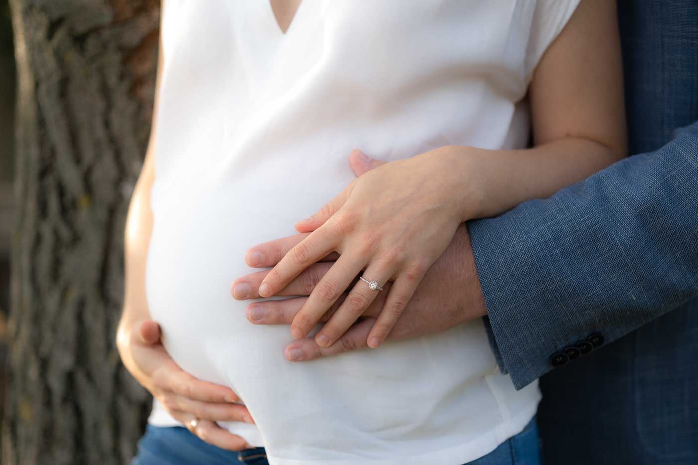 Bei einem Schwangerschaftsshooting steht eine schwangere Frau in weißer Bluse vor einem Baum, sie legt ihre mit Ehering auf die Hand ihres Mannes, der zärtlich ihren Bauch streichelt.